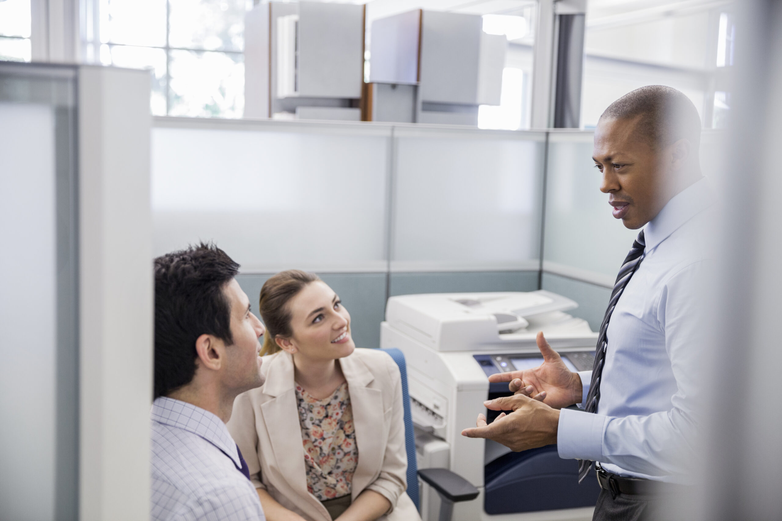 Businessman having a conversation with young professionals in office cubicle with a copier in the background signifying managed print services