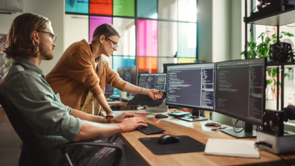 Two young professionals review code on one of three computer screens signifying Managed IT Services.