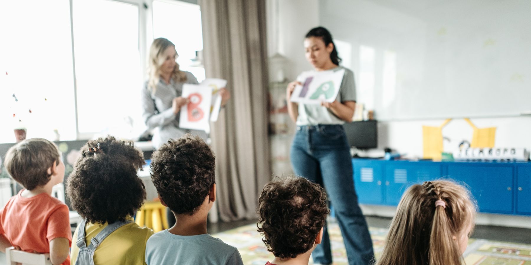 Teachers stand in front of a group of young students teaching them the letters of the alphabet.