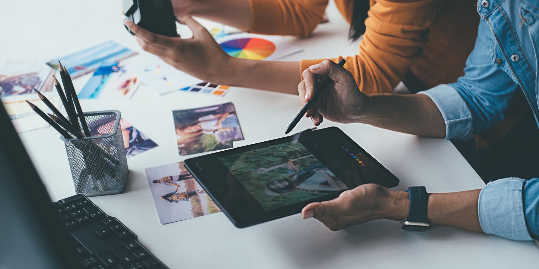 The image presents a creative professional working on a digital tablet with a stylus, indicative of a process involved in advertising design. On the tablet screen is a colorful image that the person is actively editing. Surrounding the workspace are scattered photographs, a color wheel, and pencils, suggesting a stage of conceptualizing or finalizing an advertisement. The keyboard in the foreground and the focused posture of the professional underscore the meticulous attention to detail that is characteristic of the advertising industry's creative process.