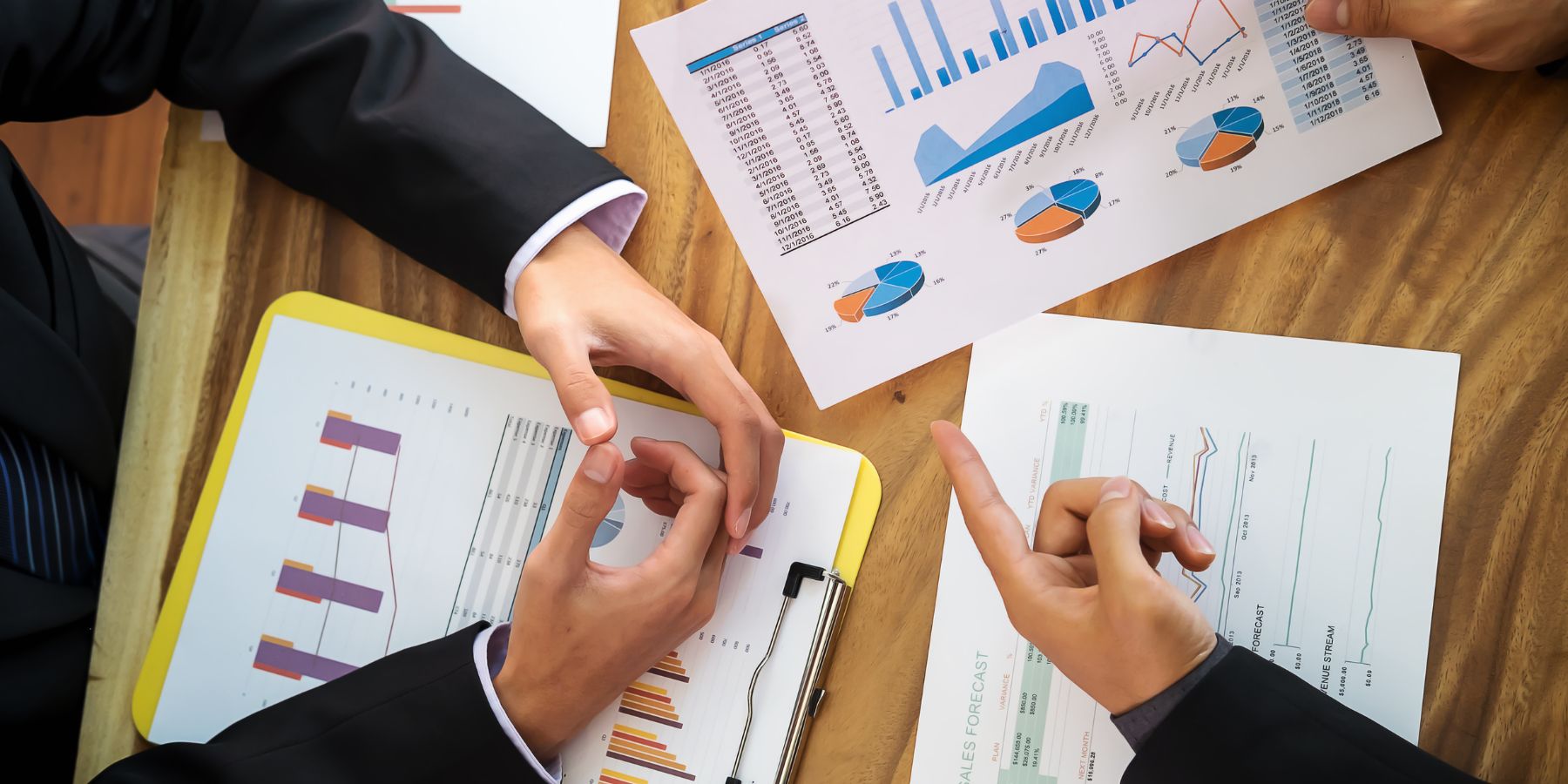 The image displays a close-up view of a financial meeting in progress, with individuals analyzing and discussing various charts and graphs. Papers are spread out on a wooden table, showing colorful pie charts, bar graphs, and data sheets that suggest a review of financial statistics or sales forecasts. The hands of the participants are visible, pointing and referencing specific data points, indicative of an in-depth financial analysis or strategic planning session. The focus on the documents conveys the importance of data-driven decision-making in the financial sector.