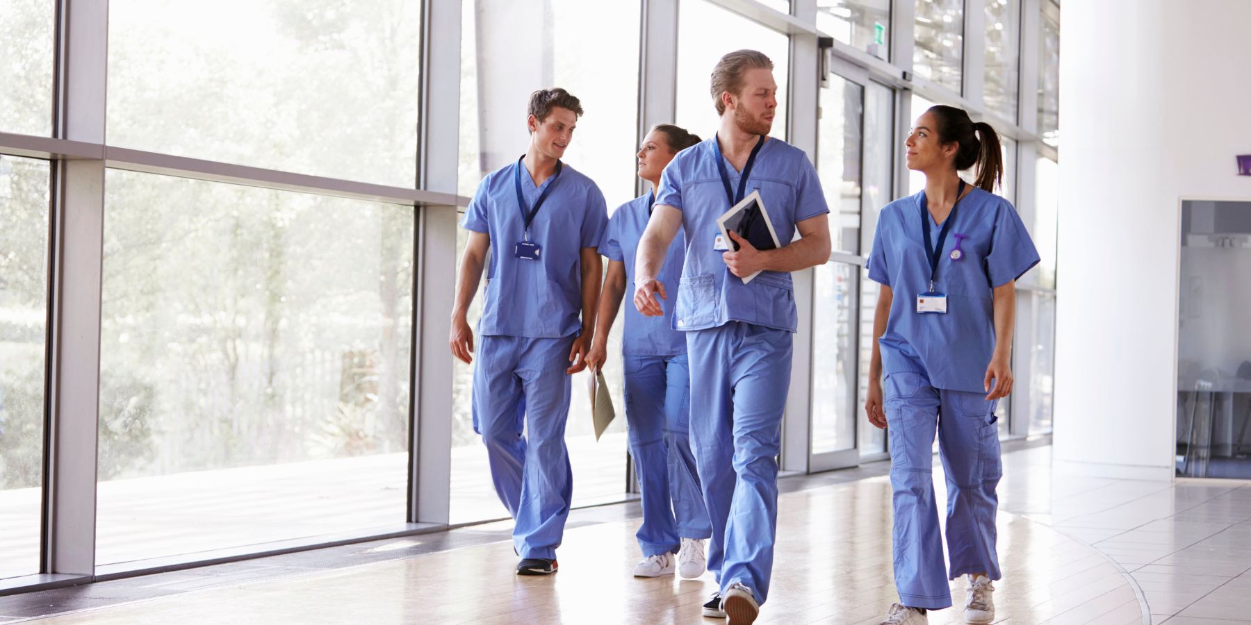 The image features a group of four healthcare professionals walking through a hospital corridor. They are dressed in blue scrubs, with ID badges visible, signifying their roles in the healthcare industry. The group appears to be in a discussion, possibly about patient care or hospital matters, as they walk past large windows that flood the space with natural light. The setting is indicative of a modern healthcare facility, emphasizing the collaborative nature of medical staff working together.