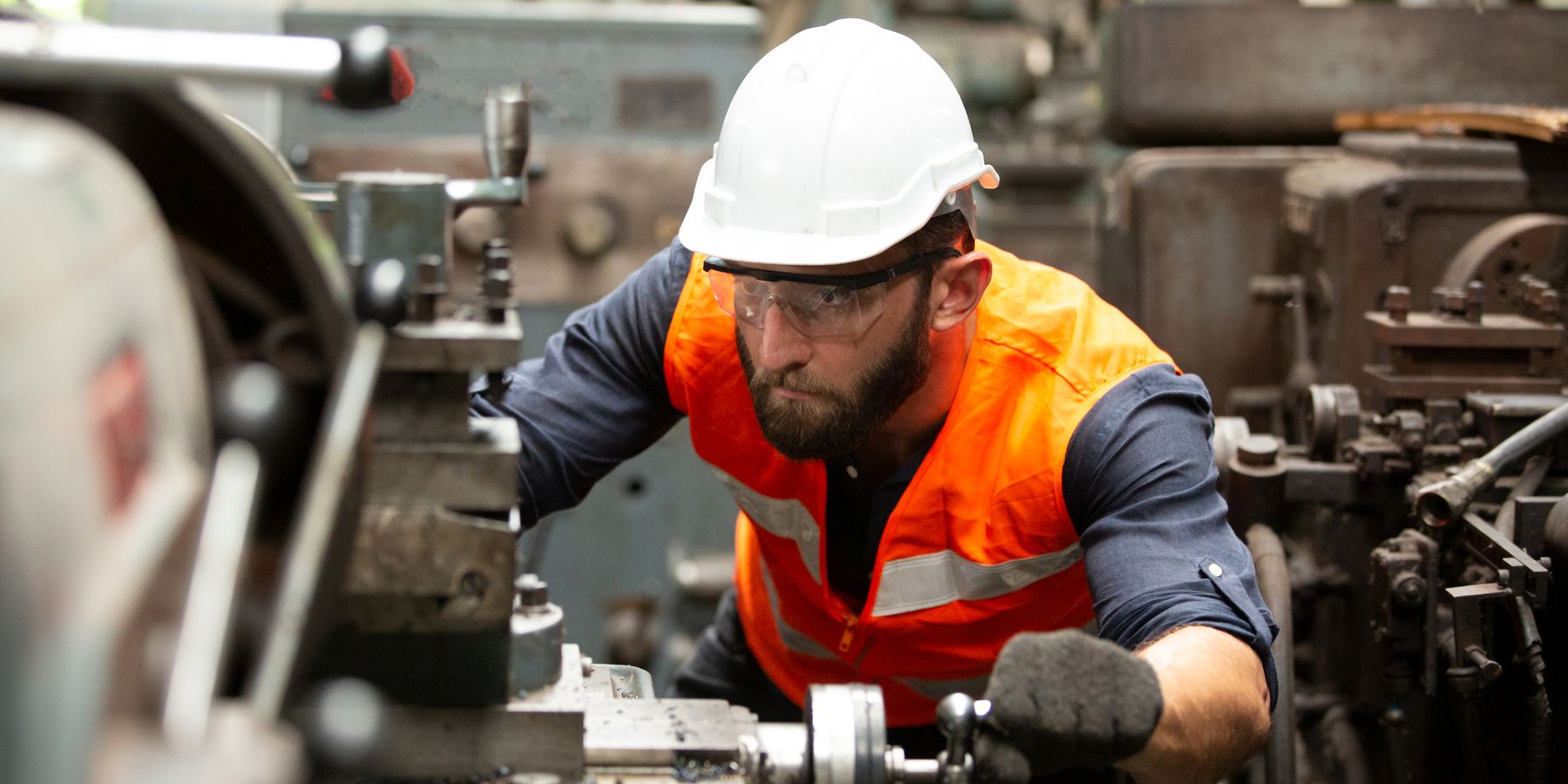 The image depicts a worker in a manufacturing setting, engaged in operating machinery. He is wearing safety gear including a white hard hat, protective eyeglasses, and a high-visibility orange vest over a long-sleeved blue shirt. His focus and hands suggest he's adjusting or inspecting the machine's components. The background is filled with industrial equipment, indicating a busy workshop environment typical in the manufacturing sector.