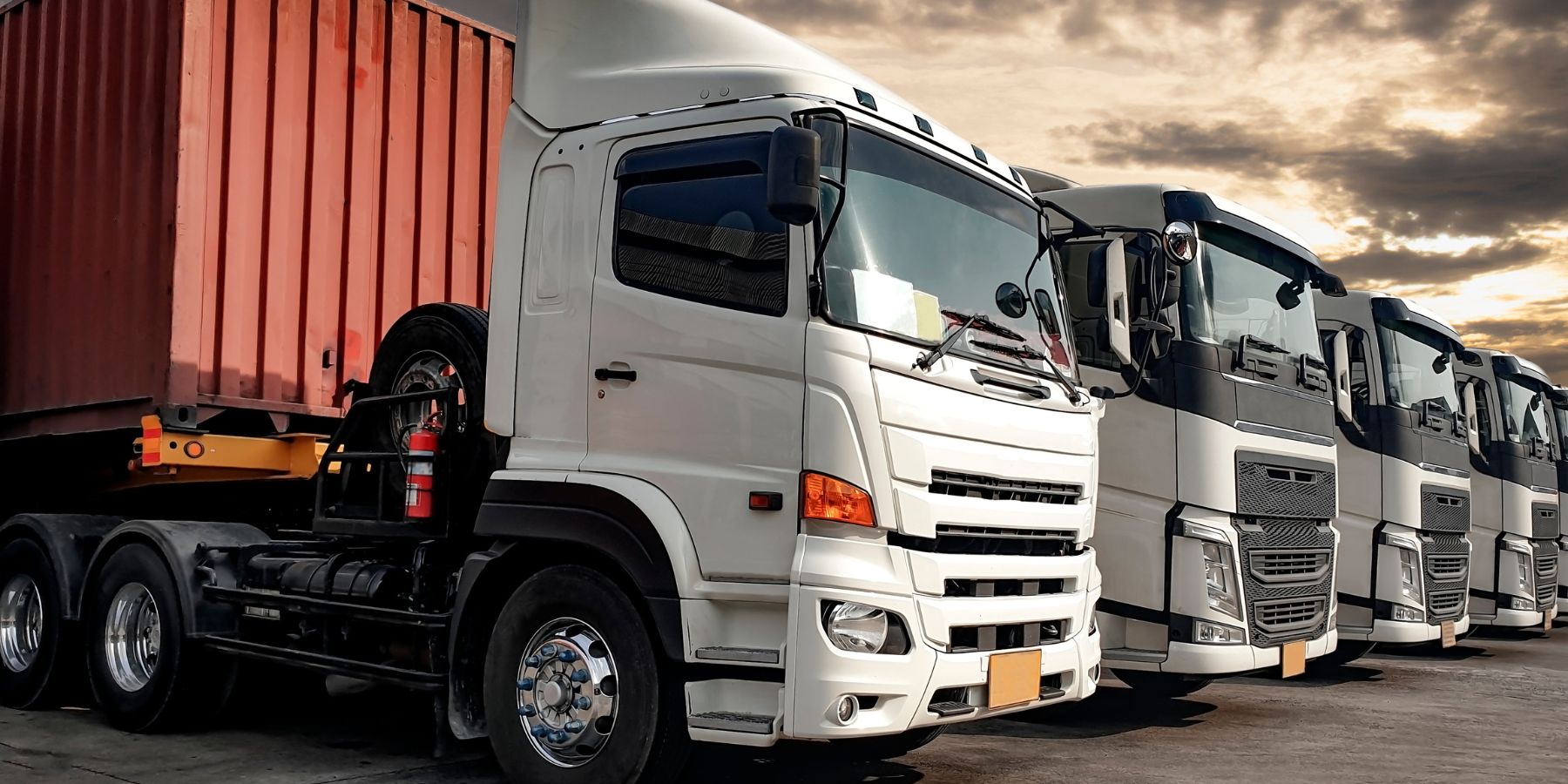 The image shows a row of modern white semi-trucks parked next to a red shipping container, indicating a scene typically associated with transportation and logistics. The trucks are aligned in an orderly fashion, showcasing their large cabs and the reflective surfaces of their clean exteriors. The setting is illuminated by a warm, ambient light suggesting either sunrise or sunset, which highlights the importance of transportation services that operate around the clock to ensure the delivery of goods.