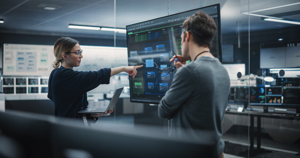 The image shows two professionals in a high-tech control room environment, engaged in a discussion about data displayed on a large monitor. The person on the left, a woman with glasses, is pointing at a specific section of a graphical user interface on the screen, which appears to show a complex system or network diagram. The person on the right, a man holding a laptop, is looking closely at the point of interest, suggesting a collaborative analysis or problem-solving session. The room is equipped with multiple screens and monitors, indicating a sophisticated setup that is typical for a Managed IT Services provider. The setting conveys a sense of advanced technical management, with a focus on monitoring and optimizing IT systems for efficiency and reliability.