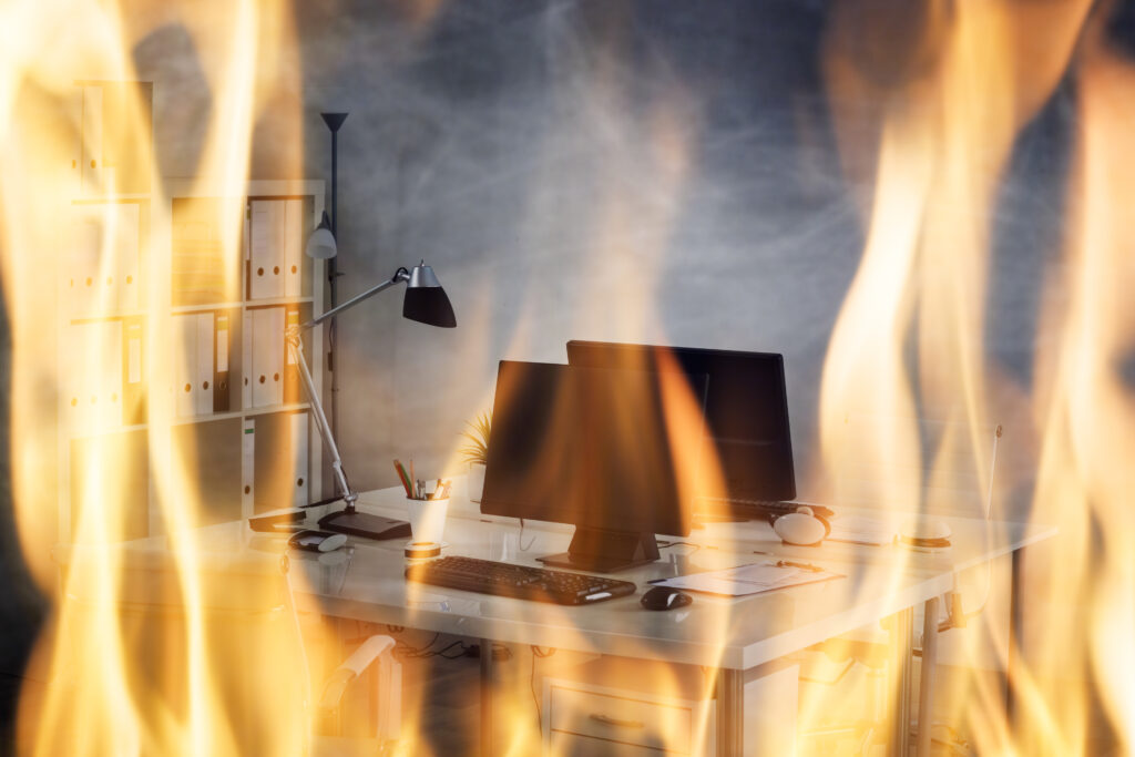 An office desk and computer engulfed in flames, symbolizing the critical need for business continuity planning in the event of a disaster. The desk is well-equipped with a monitor, keyboard, mouse, and desk lamp, all shrouded in fire, highlighting the importance of having a plan to protect and recover business operations.
