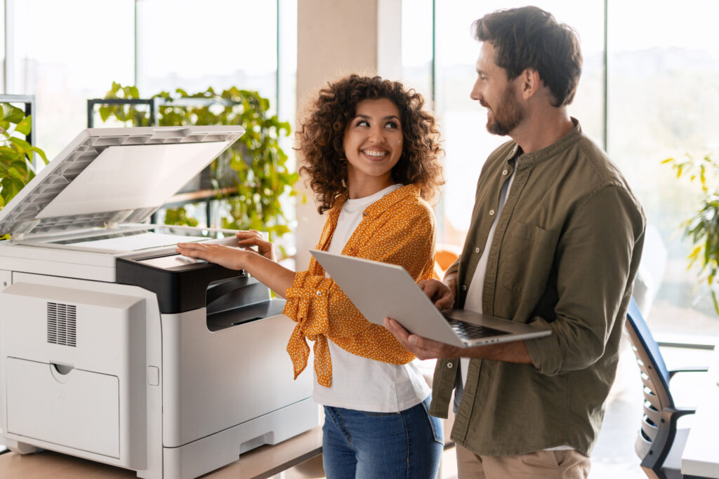 Two office employees using a multifunction printer and laptop in a modern workspace, illustrating printer management for efficient business printing workflows.